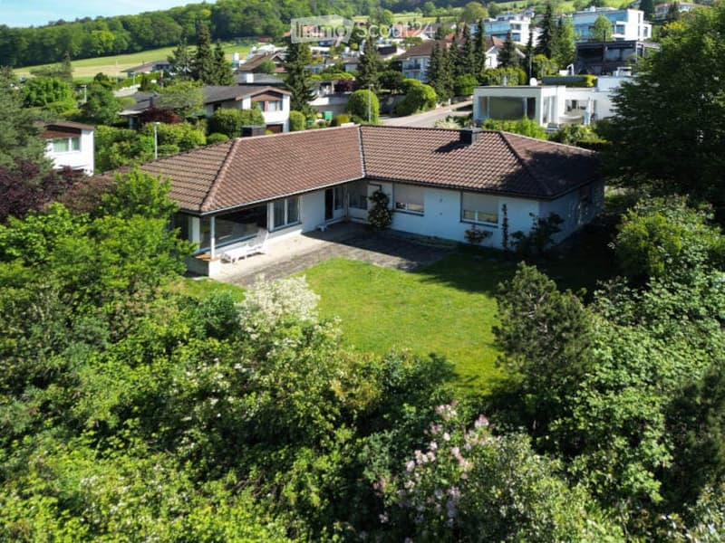 Aerial view of a two-story house with a tiled roof, surrounded by a well-maintained lawn and trees, with neighboring houses visible in the background.