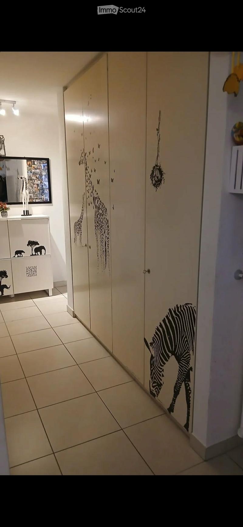 A narrow hallway with tiled flooring, white walls, and decorative animal wall decals including giraffes, zebras, and elephants. A small cabinet with a mirror and a small plant is visible on the left side.