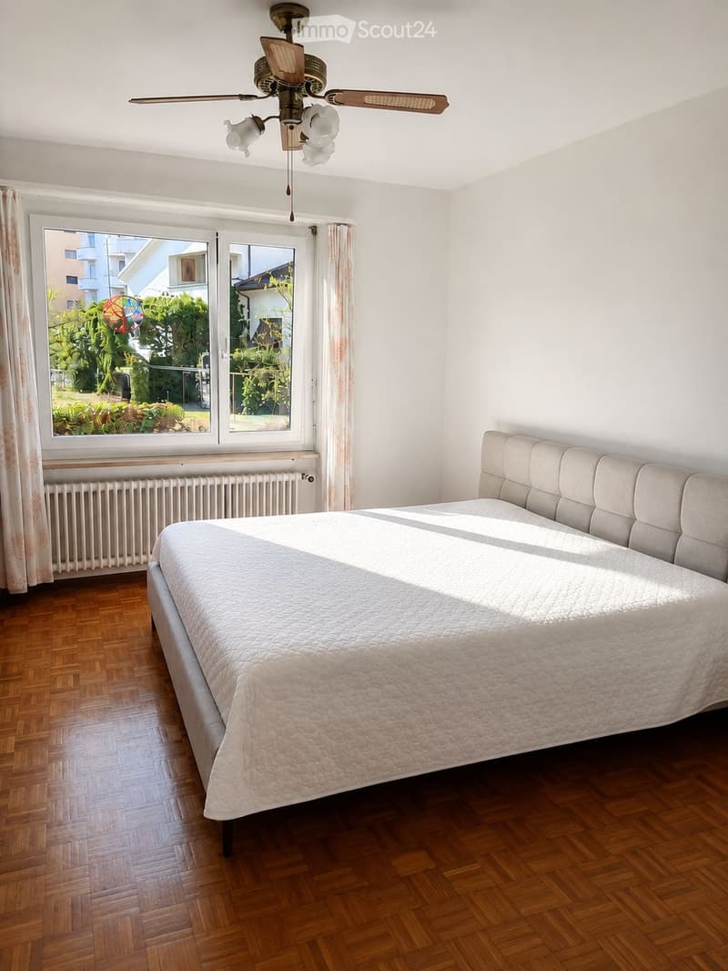 Bright bedroom with wooden flooring, large window with curtains, a bed with a headboard, and a ceiling fan with light fixtures.