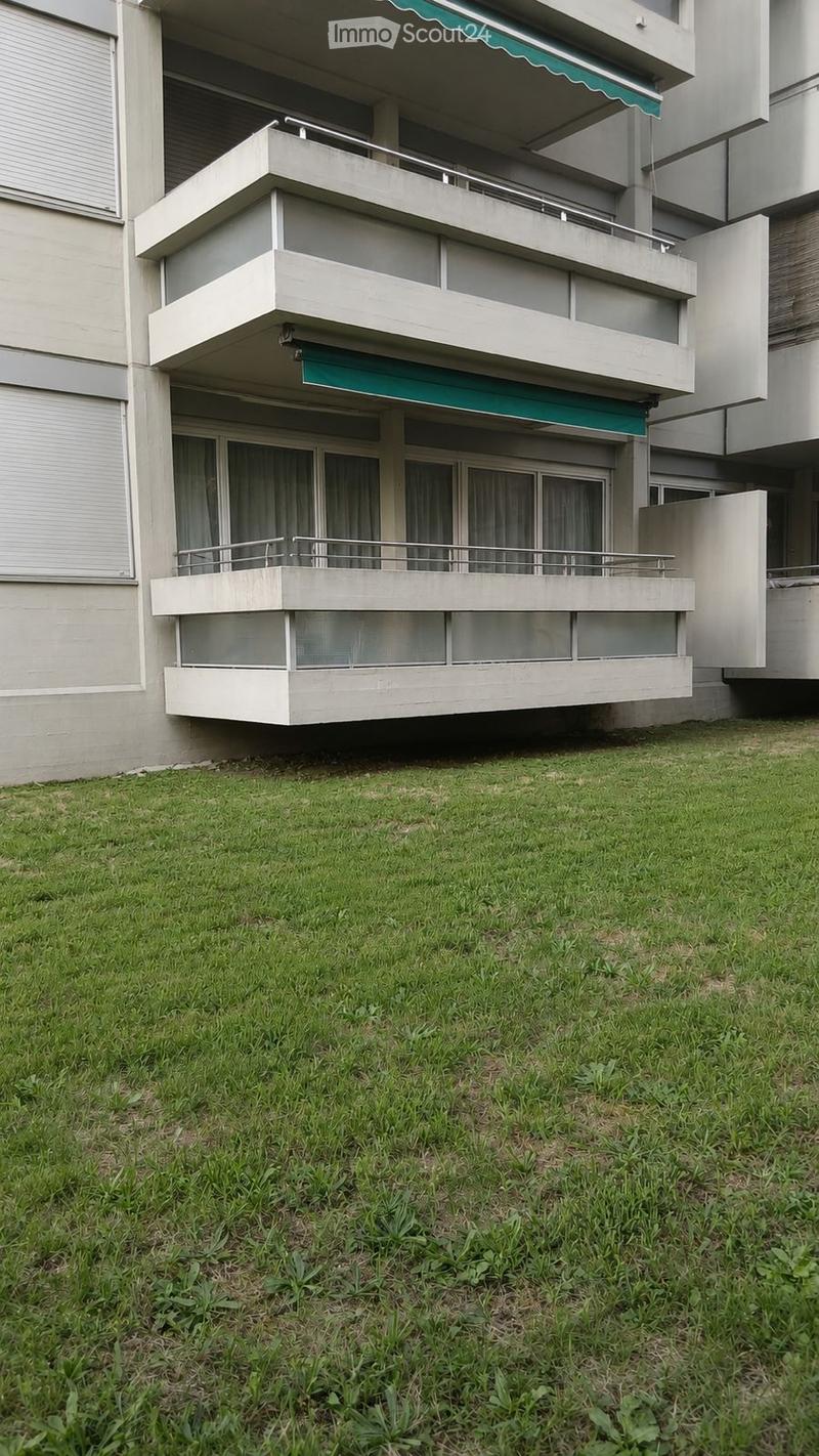 Multi-story building with balconies and green awnings, situated on a raised ground floor with a grassy area in front.