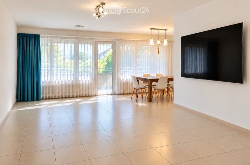Bright dining area with a wooden dining table, white chairs, large windows with sheer curtains, and a flat-screen TV mounted on the wall.