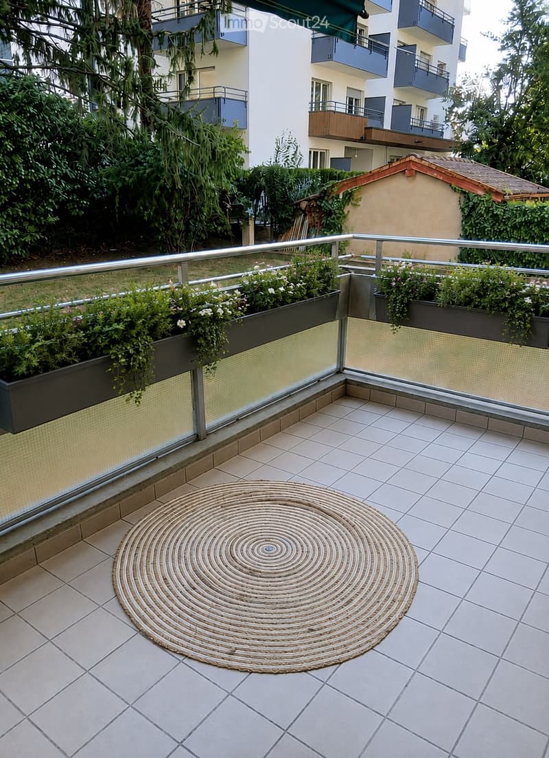 Balcony with tiled floor, round woven rug, glass railing, and potted plants.
