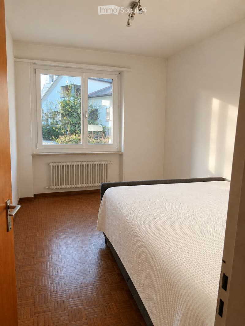 Bright bedroom with wooden parquet flooring, a single bed with white bedding, a radiator below a large window, and modern ceiling lights.