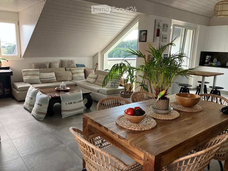 A modern, well-lit living and dining area with a large sectional sofa, coffee table, dining table with chairs, and decorative bowls. The space features large windows with a scenic view, potted plants, and a sleek kitchen area in the background.