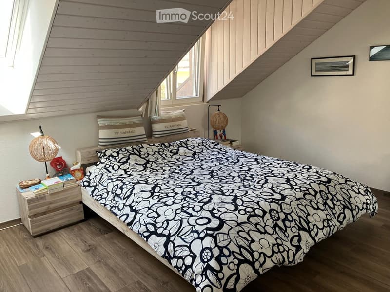 Cozy bedroom with wooden floors, slanted ceiling, large window, and a bed with a black and white floral duvet.