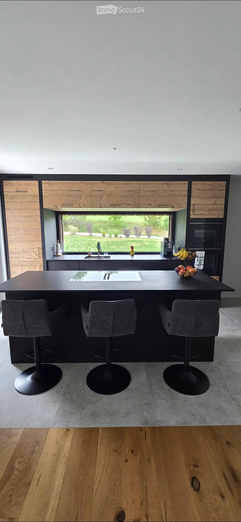 Modern kitchen with a central island, three stools, a sink, and a view of a garden through large windows.