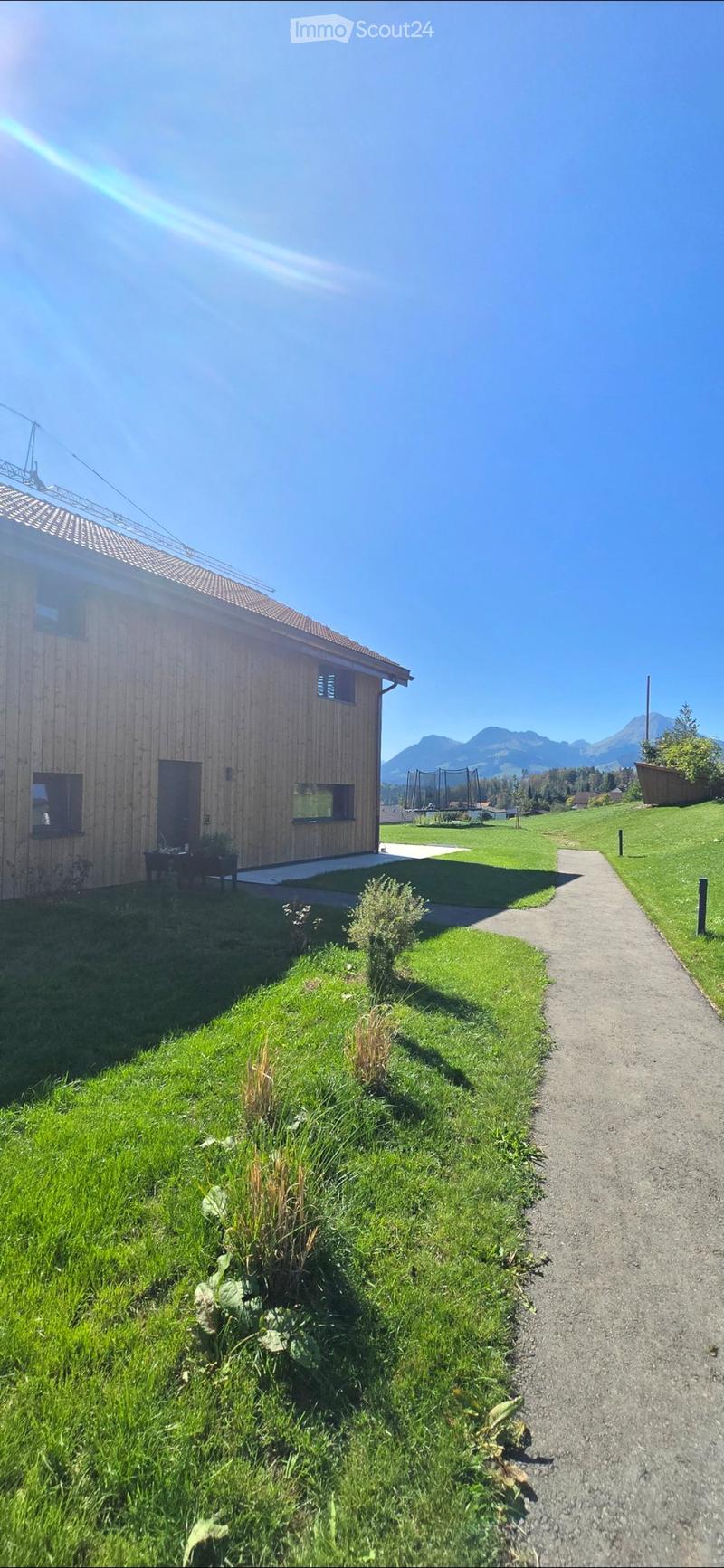 A modern wooden house with a sloped roof, surrounded by a well-maintained lawn and a gravel pathway leading to the entrance. The background features a scenic mountain view and a fenced play area.