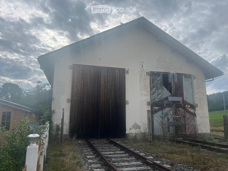 A small, white, single-story building with a pitched roof and weathered wooden doors, situated near a railway track.