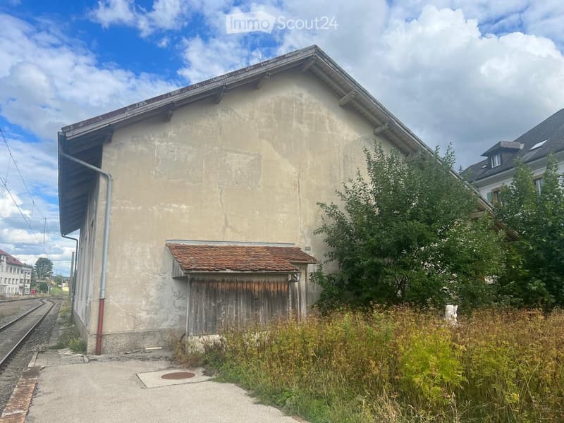 Two-story building with beige walls, a slanted roof, and a small wooden shed attached to the side. The building is adjacent to railway tracks and has overgrown vegetation in front.