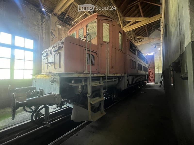 An old train displayed inside a large, dimly lit workshop with wooden beams and large windows. The train is mounted on railway tracks.