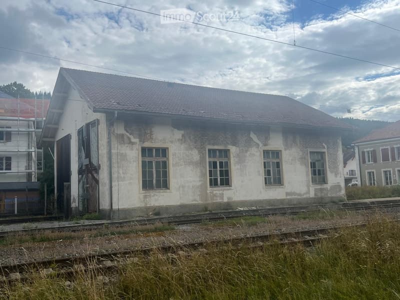 Two-story building with a tiled roof, brick facade, and multiple windows. The building appears to be in a rural setting with railway tracks in the foreground.