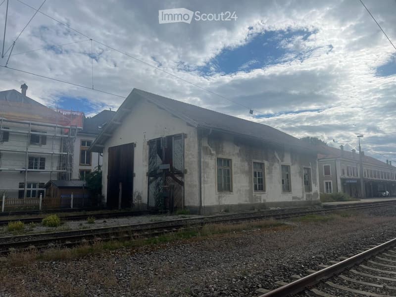 Old, weathered building with multiple windows and a large wooden door, situated near railway tracks.