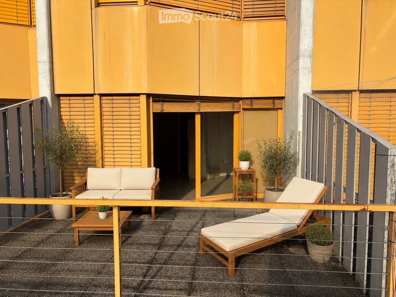 Balcony with a white sofa, wooden sun lounger, small wooden tables, and potted plants. The balcony has a modern design with wooden blinds and a metal railing.