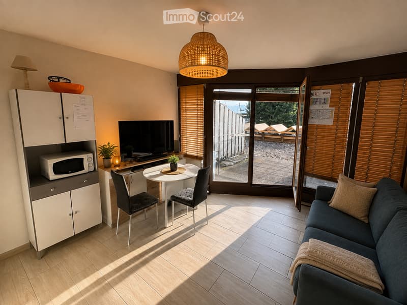Bright, modern living space with a sofa, coffee table, dining table, chairs, microwave, TV, and a cabinet with a lamp on top.