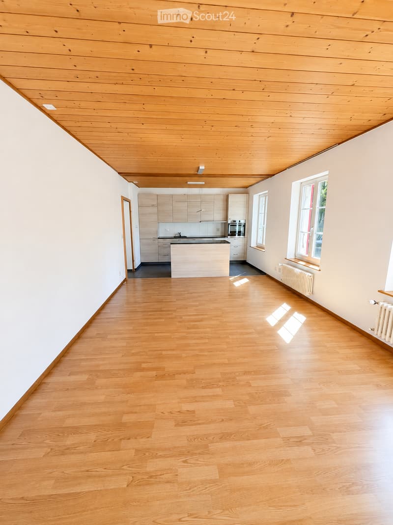 Empty room with light wooden flooring, white walls, and a modern kitchen area in the background. The space features large windows allowing natural light to fill the room.