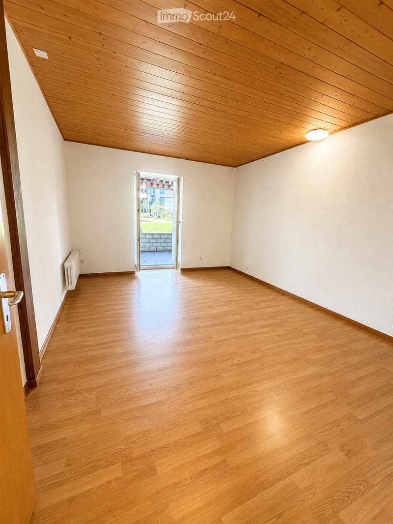 Empty room with wooden flooring, white walls, and a wooden ceiling. A door leads to an outdoor area with a view of greenery.