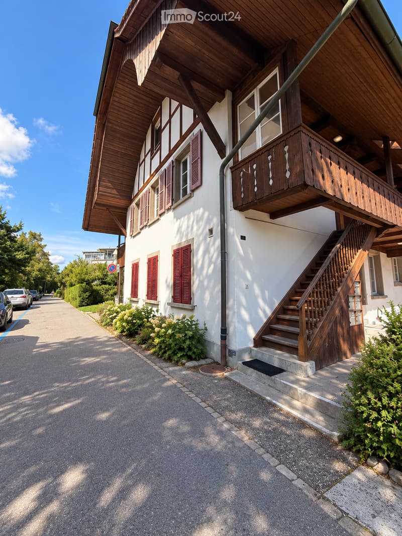 Two-story house with white walls, wooden roof, red shutters, and a wooden balcony with railings. Stairs lead up to the entrance.
