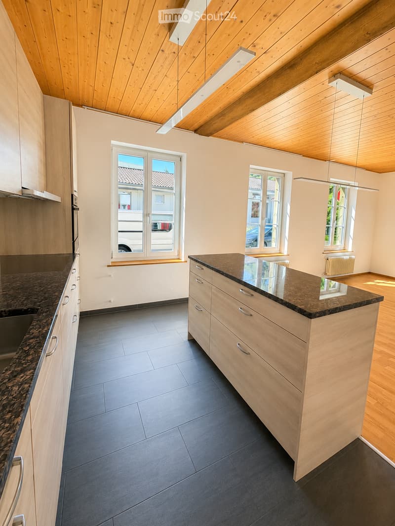 Modern kitchen with light wooden cabinets, dark granite countertops, and dark tiled flooring. Features include a central island, built-in sink, and large windows with views of the street.