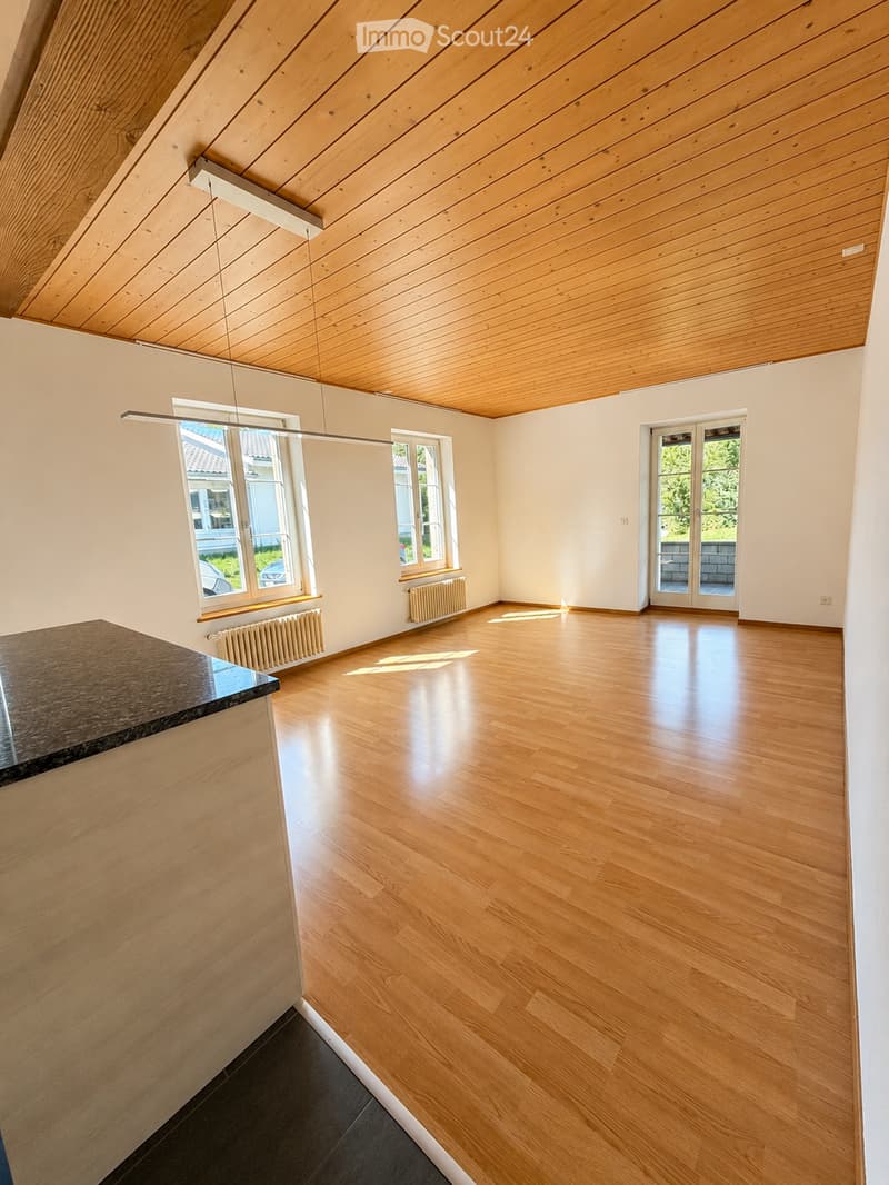 Bright, empty room with wooden flooring, white walls, and large windows. Features a wooden ceiling and a sliding glass door leading outside.