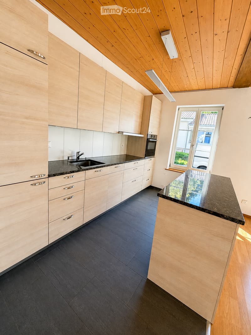 Modern kitchen with light wooden cabinets, stainless steel sink, oven, and a central island with a dark countertop.