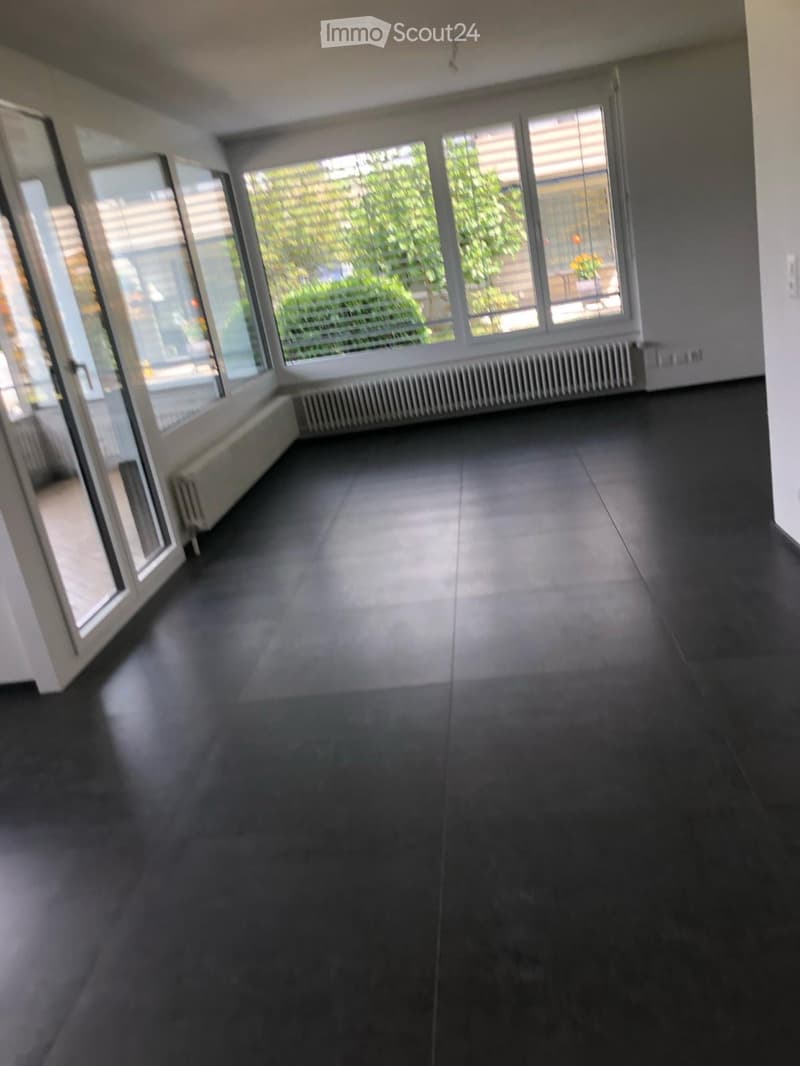 Empty room with dark tiled flooring, large windows with blinds, and radiators along the walls.