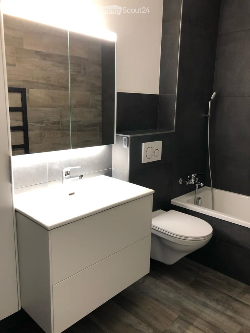 Modern bathroom with a white sink, toilet, bathtub, and a large mirror with built-in lighting. Dark tiled walls and wooden flooring.