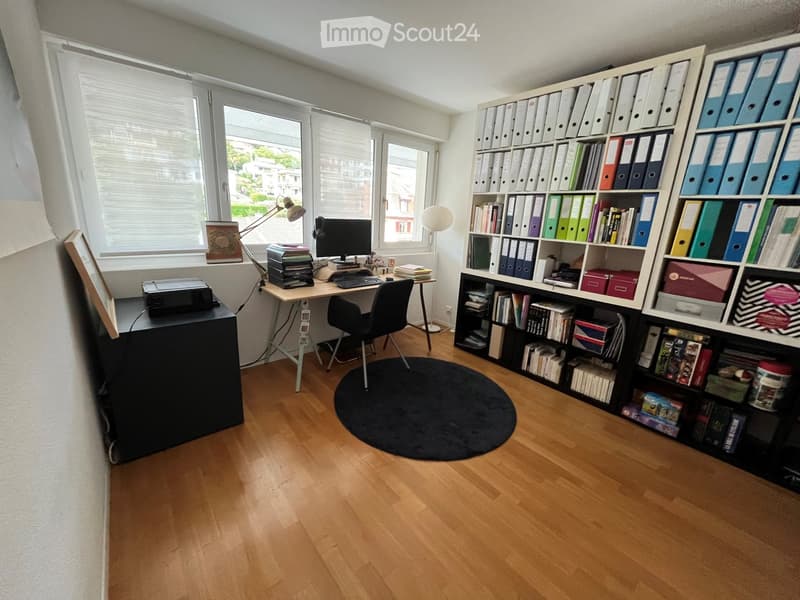 Well-lit study or office space with wooden flooring, large windows, a desk with a computer, chair, printer, and a bookshelf filled with books and file folders.