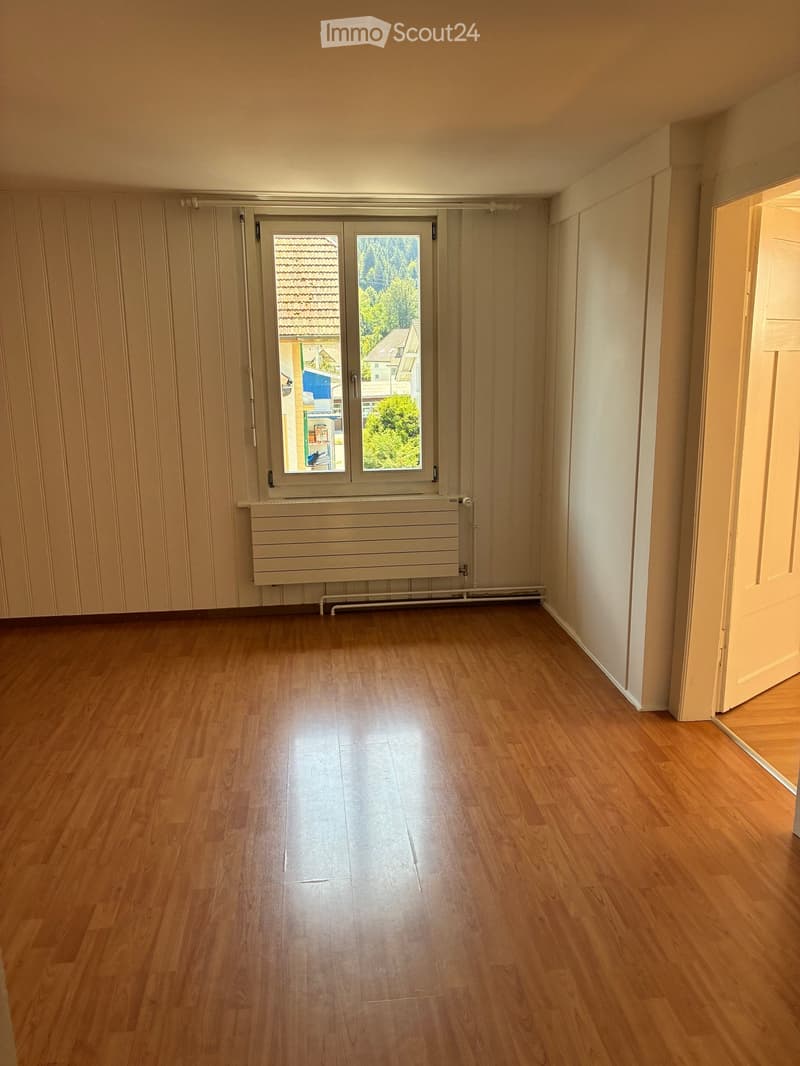 Empty room with wooden flooring, white walls, and a large window with a radiator below it. The room has a door on the right side.