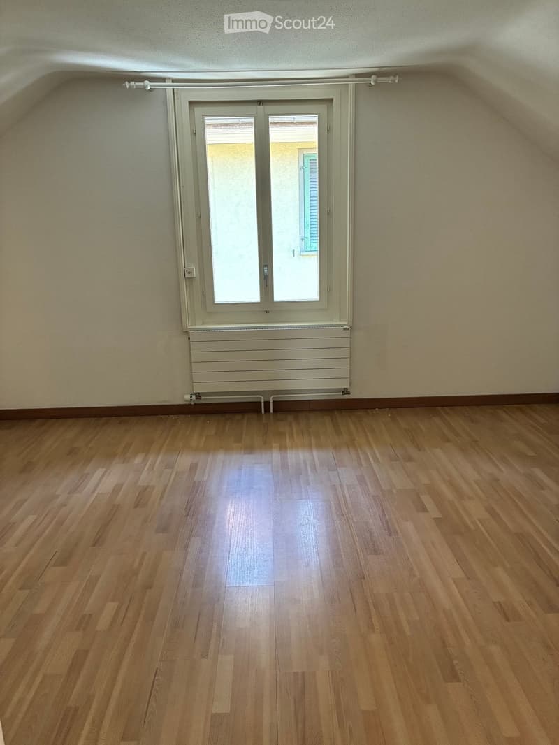 Empty room with wooden flooring, white walls, and a large window with blinds. The room has a radiator below the window and a simple ceiling with visible beams.