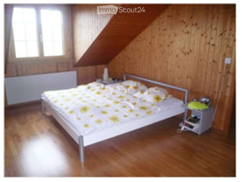 Cozy attic bedroom with wooden walls and floor, large window, single bed with yellow and white bedding, nightstand with lamp.