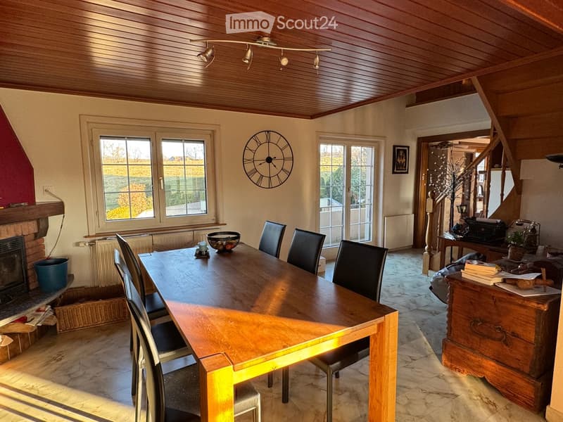 Dining room with wooden dining table, chairs, fireplace, large windows, and a clock on the wall.