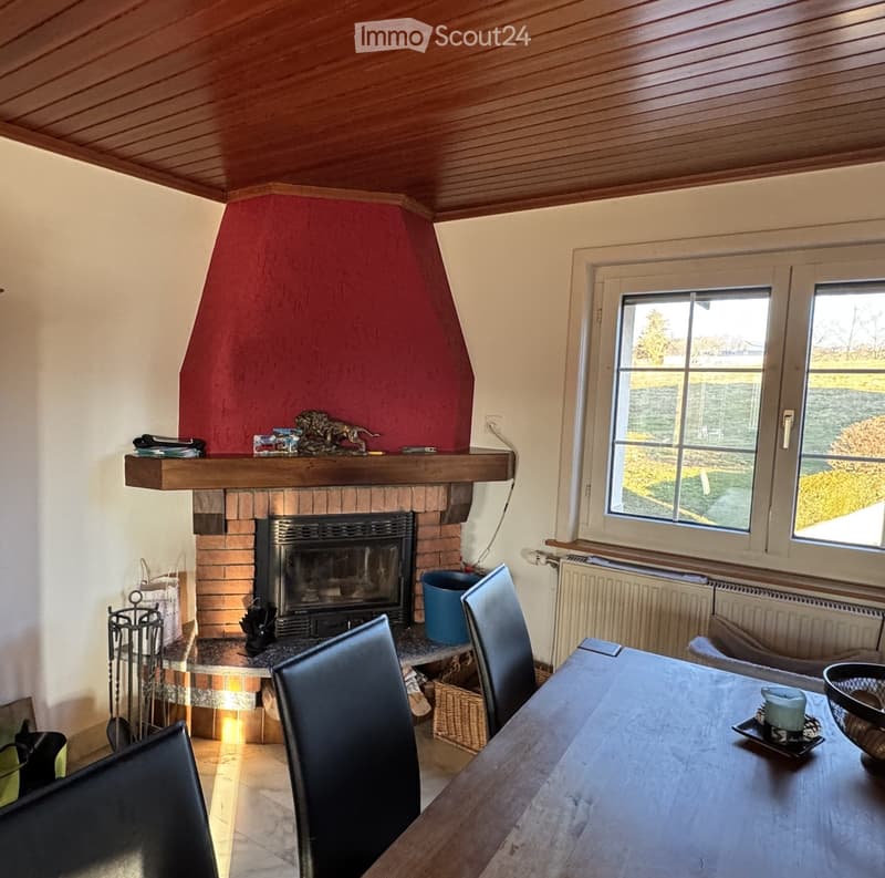 Cozy living room with a brick fireplace, wooden table, and chairs, featuring a large window with a view of the garden.