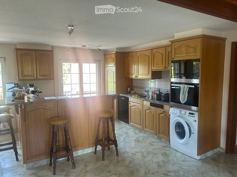 Bright kitchen with wooden cabinets, marble flooring, and three wooden stools at the island. Appliances include a washing machine, dishwasher, oven, microwave, and coffee machine.