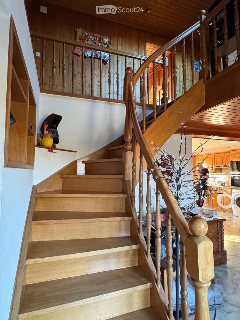 Wooden staircase with a handrail, leading to an upper level. The walls are painted white, and there are decorative items on a shelf to the left. The lower level shows a glimpse of a kitchen area with cabinets and appliances.