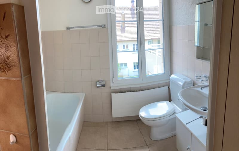 Bathroom with a bathtub, toilet, sink, mirror, and radiator. The room has tiled walls and a window offering a view of the roof and surrounding buildings.