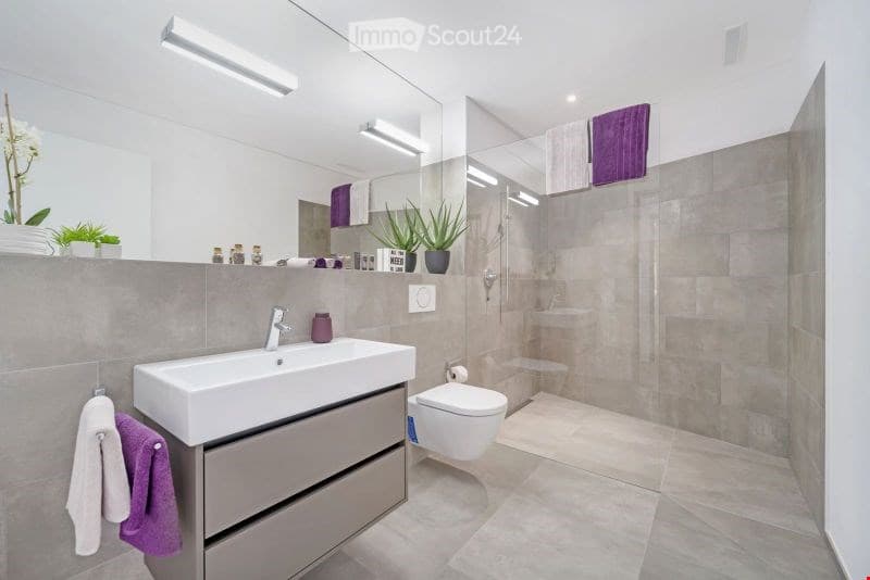 Modern bathroom with a white sink, large mirror, toilet, and glass-enclosed shower. Features grey tiles, purple towels, and decorative plants.