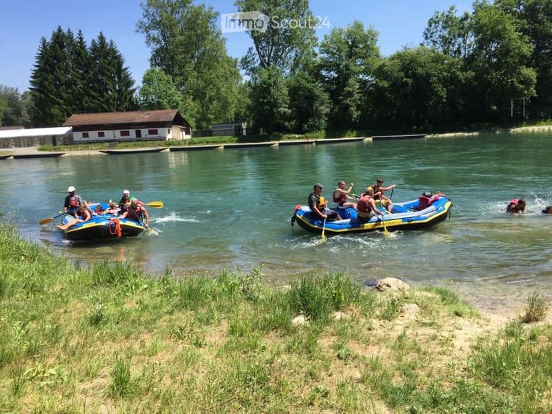 People in inflatable rafts on a calm river, surrounded by trees and a small building on the shore.