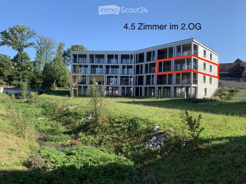 Modern multi-story apartment building with balconies, surrounded by green space and a small stream.