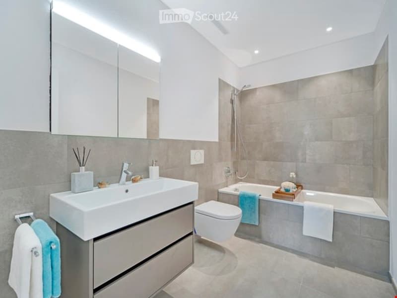 Modern bathroom with a white sink, chrome faucet, large mirror, toilet, bathtub, and towels neatly placed on the edge of the tub.