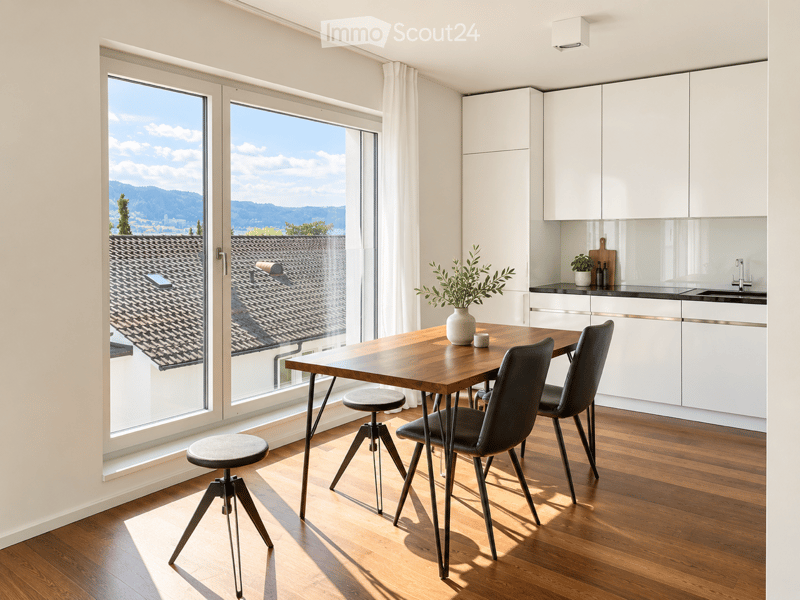 Modern kitchen and dining area with large windows offering a view of a tiled roof and distant landscape. Features a wooden dining table with four chairs, two stools, white kitchen cabinets, a sink, and a vase with a plant on the table.