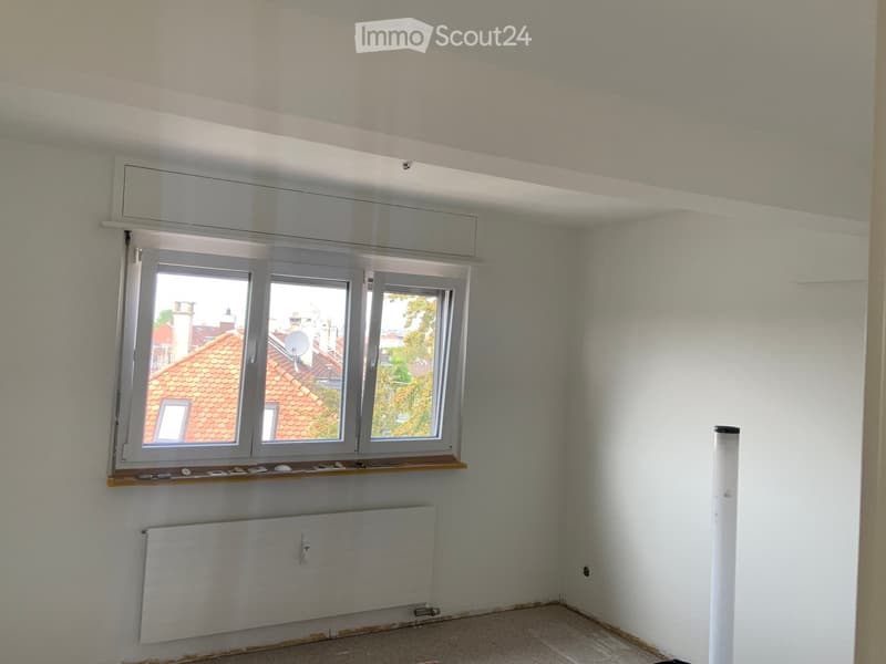 Empty room with white walls, large window with wooden sill, radiator, and visible electrical outlets.