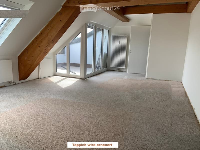 Empty attic room with exposed wooden beams, large windows, and carpeted floor.