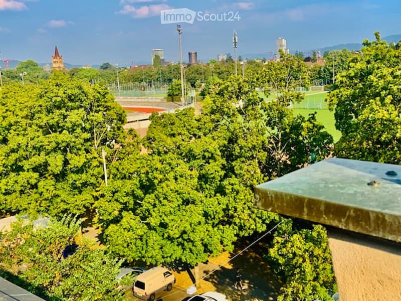Aerial view of a balcony overlooking a lush green park area with trees, a sports field, and distant cityscape with buildings and towers.