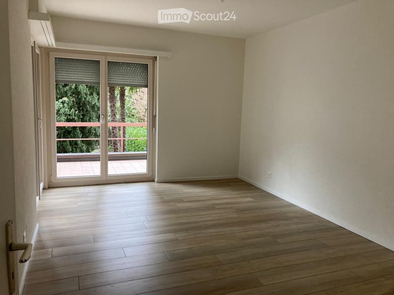Empty room with wooden flooring, light-colored walls, and a large sliding glass door leading to a balcony with outdoor furniture visible.