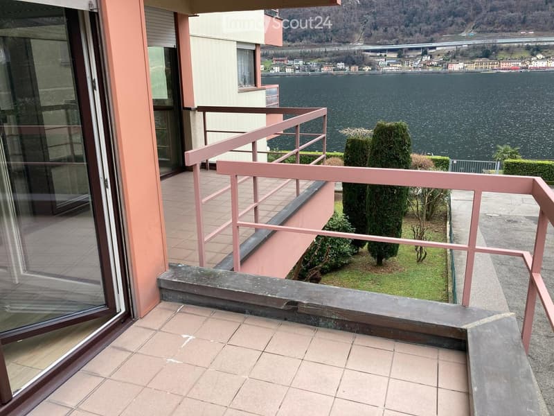 Balcony with pink railing, tiled floor, overlooking a lake and town, with small garden area and trimmed trees.