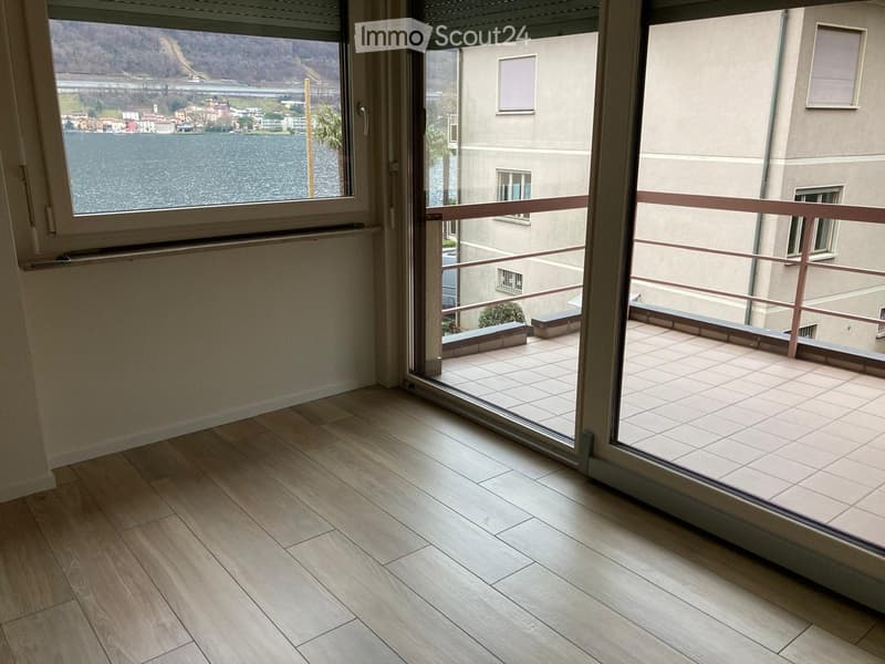 Balcony with wooden flooring, glass sliding doors, and a view of a lake and surrounding buildings.