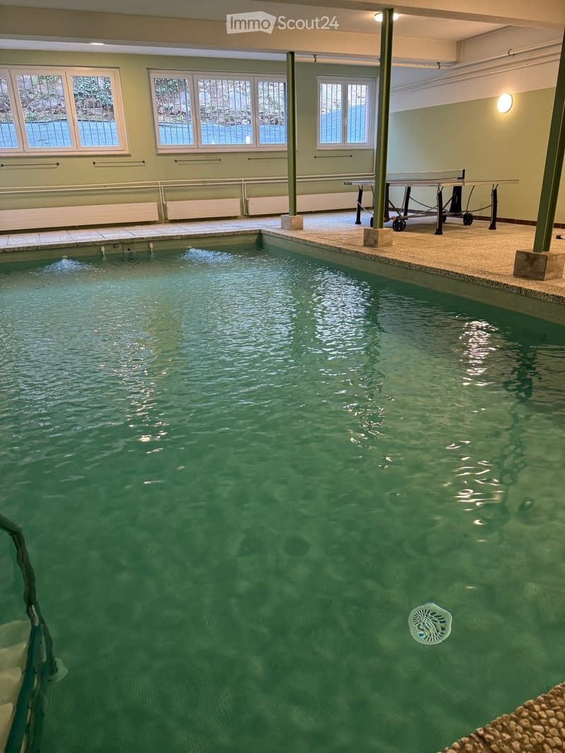 Indoor swimming pool area with clear water, surrounded by windows and railings, with a small table and chairs nearby.