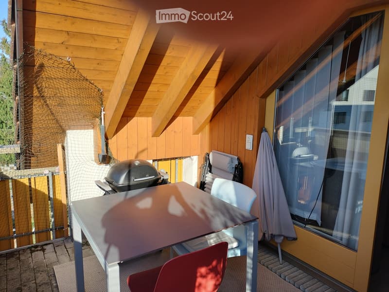 Balcony with wooden ceiling, glass window, table, chairs, umbrella, and grill.