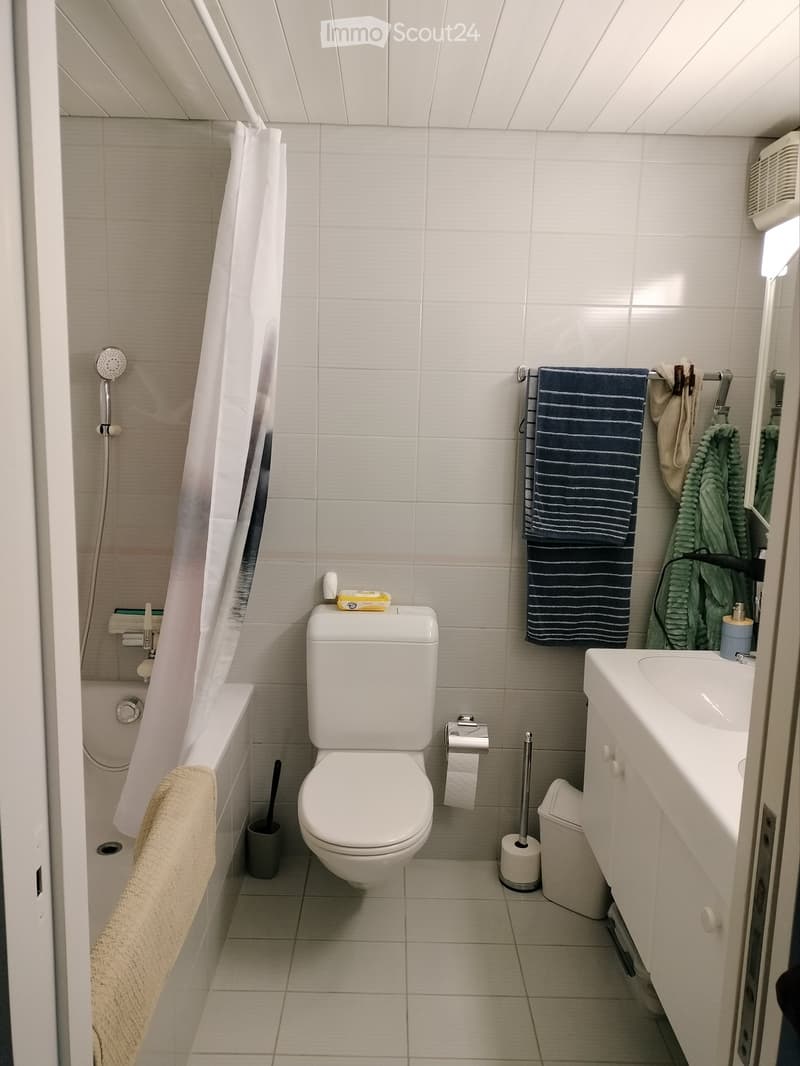 Modern bathroom with white tiles, a toilet, a sink, a bathtub, and towels hanging on a rack. The shower area is enclosed with a white curtain, and a small trash bin is placed near the toilet.