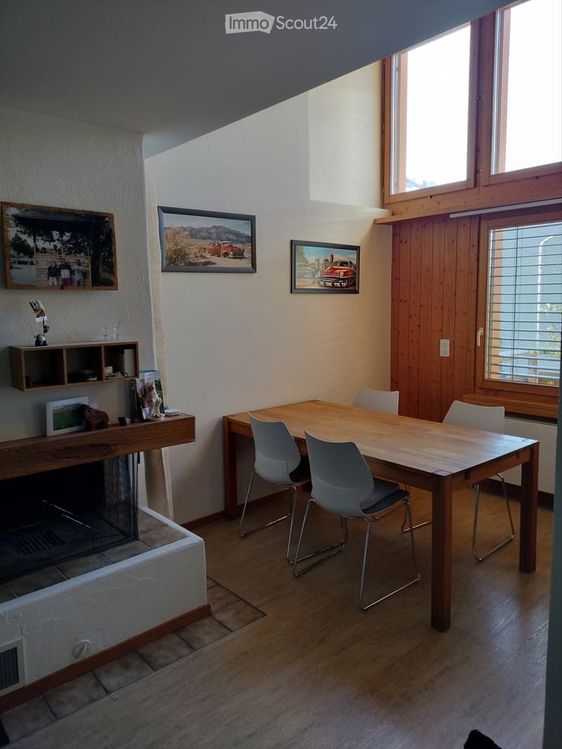 Bright room with wooden flooring, a central dining table with four modern chairs, a fireplace with a wooden shelf above it, and several picture frames on the walls.
